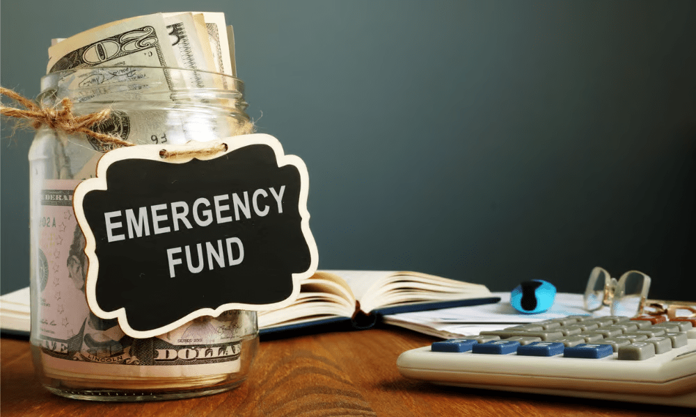 jar filled with cash on a desk with a calculator, book and glasses with a sign that reads Emergency Fund